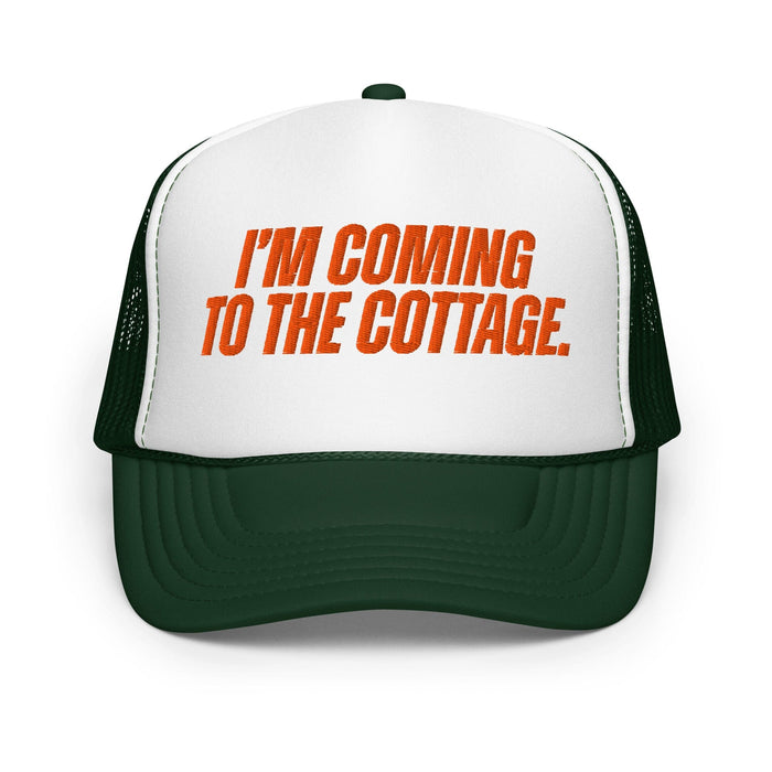 A dark green and white trucker hat with orange text on the front panel that reads 'I'M COMING TO THE COTTAGE.'