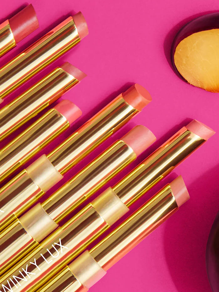 eight nude shades of skinny plump lipstick flat lay on pink background with plum